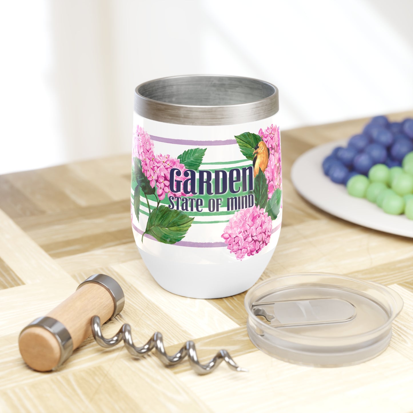 Garden State illustrated wine tumbler with hydrangeas and bird and green and purple stripe next to plastic lid with closure on cutting board with wine opener and plate of grapes