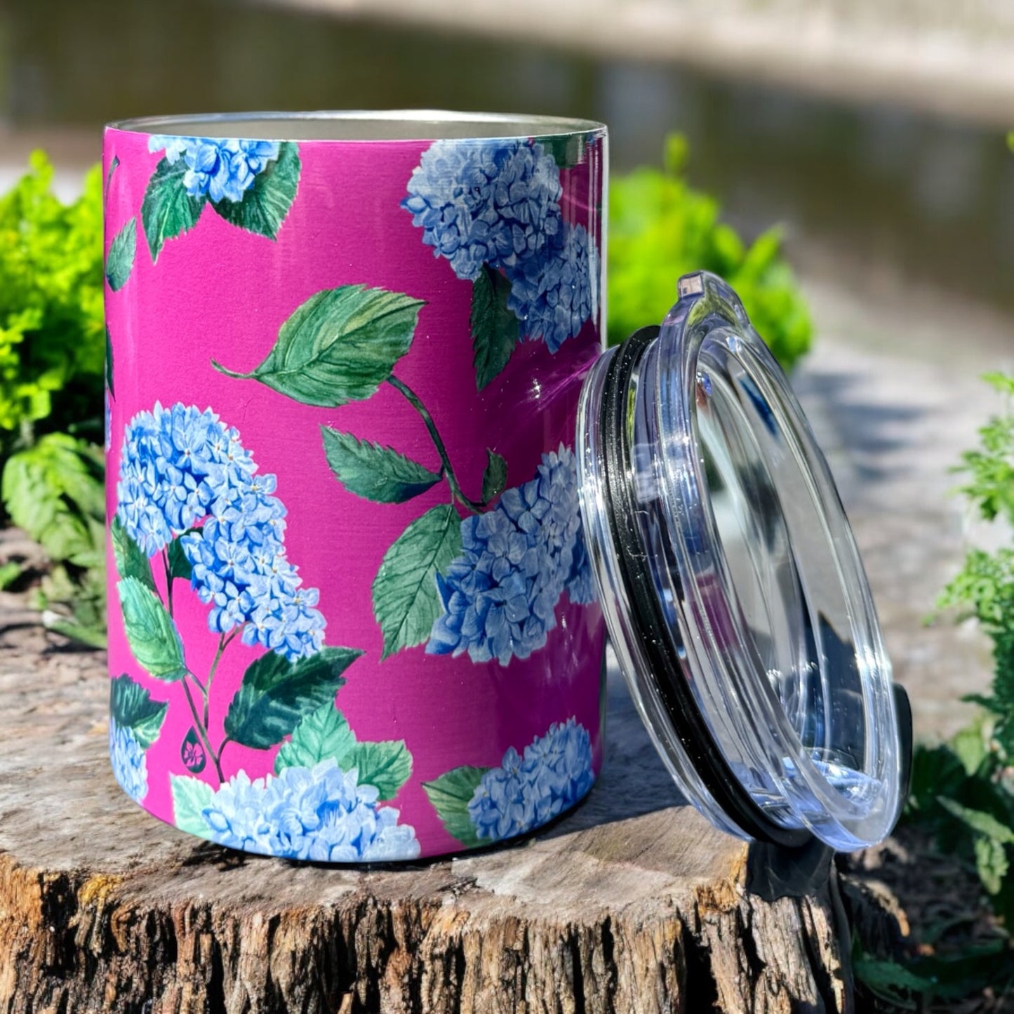 Hydrangea Tumbler in Peony sitting on a tree stump with clear lid leaning against Tumbler