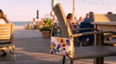 A cream shoulder bag with a colorful butterfly and floral print hangs from a woven outdoor chair on a wooden boardwalk at sunset, with beachside seating and people blurred in the background.