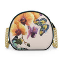 A round shoulder bag with a black leather trim and a gold chain strap, featuring a colorful print of yellow orchids and blue, green, and yellow butterflies on a light beige background.