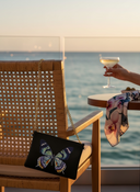 The Solstice Bag hanging off a wicker restaurant chair with a luxury ocean view and woman drinking a cocktail. 
